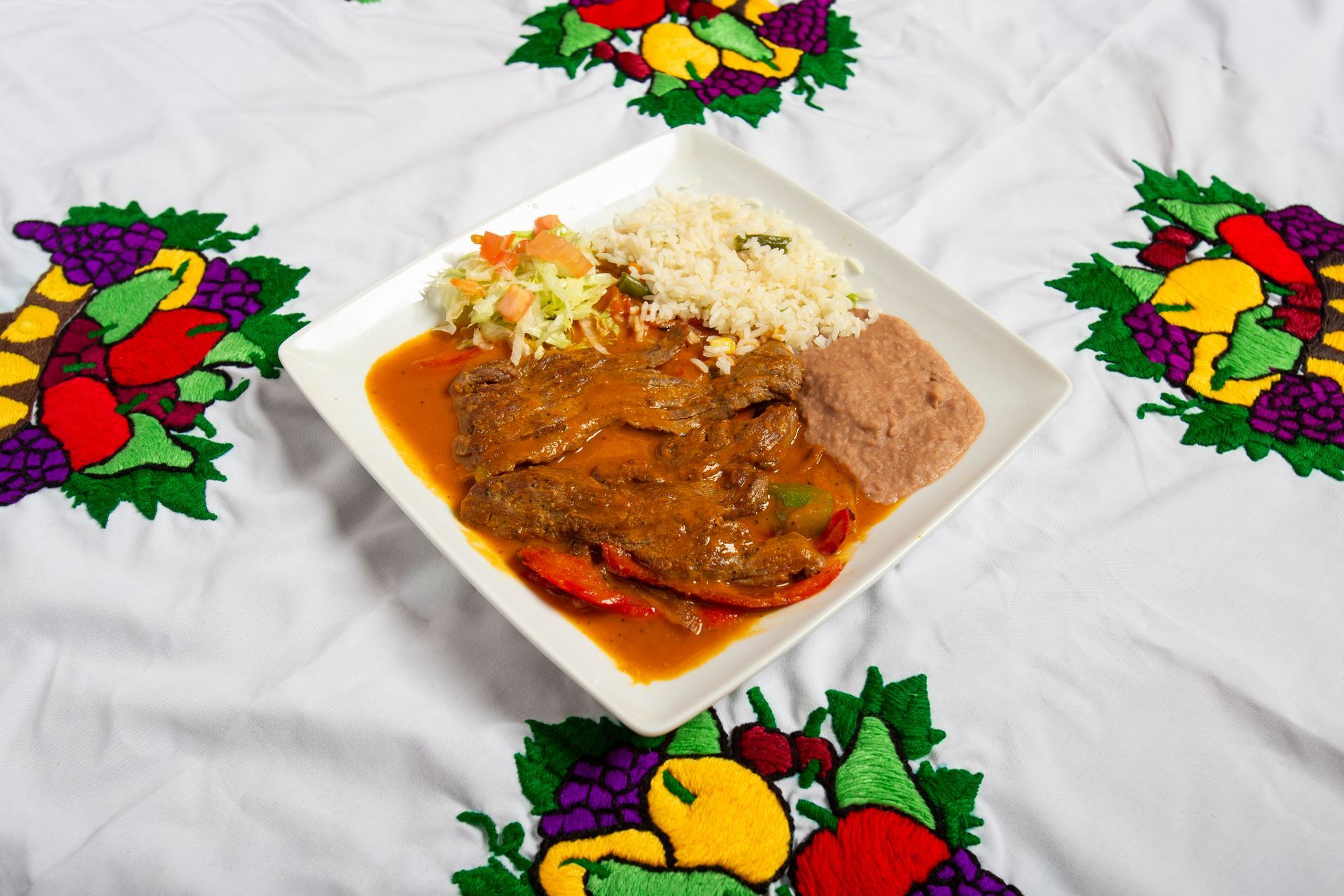 Plate with meat in sauce, rice, beans, and salad on a white tablecloth with fruit embroidery.