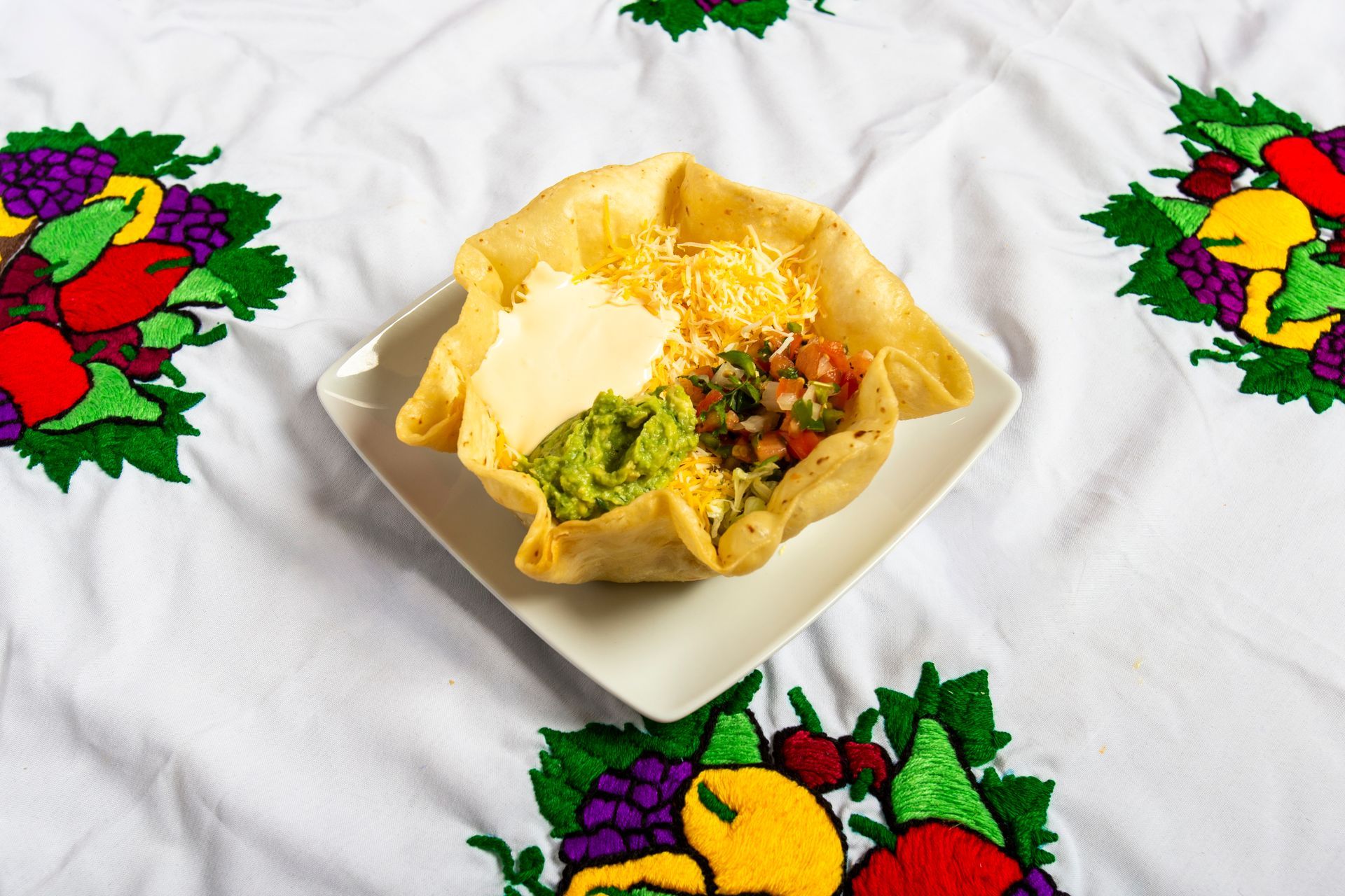 Taco salad in a fried tortilla bowl on a white square plate, with guacamole, salsa, cheese, and sour cream.