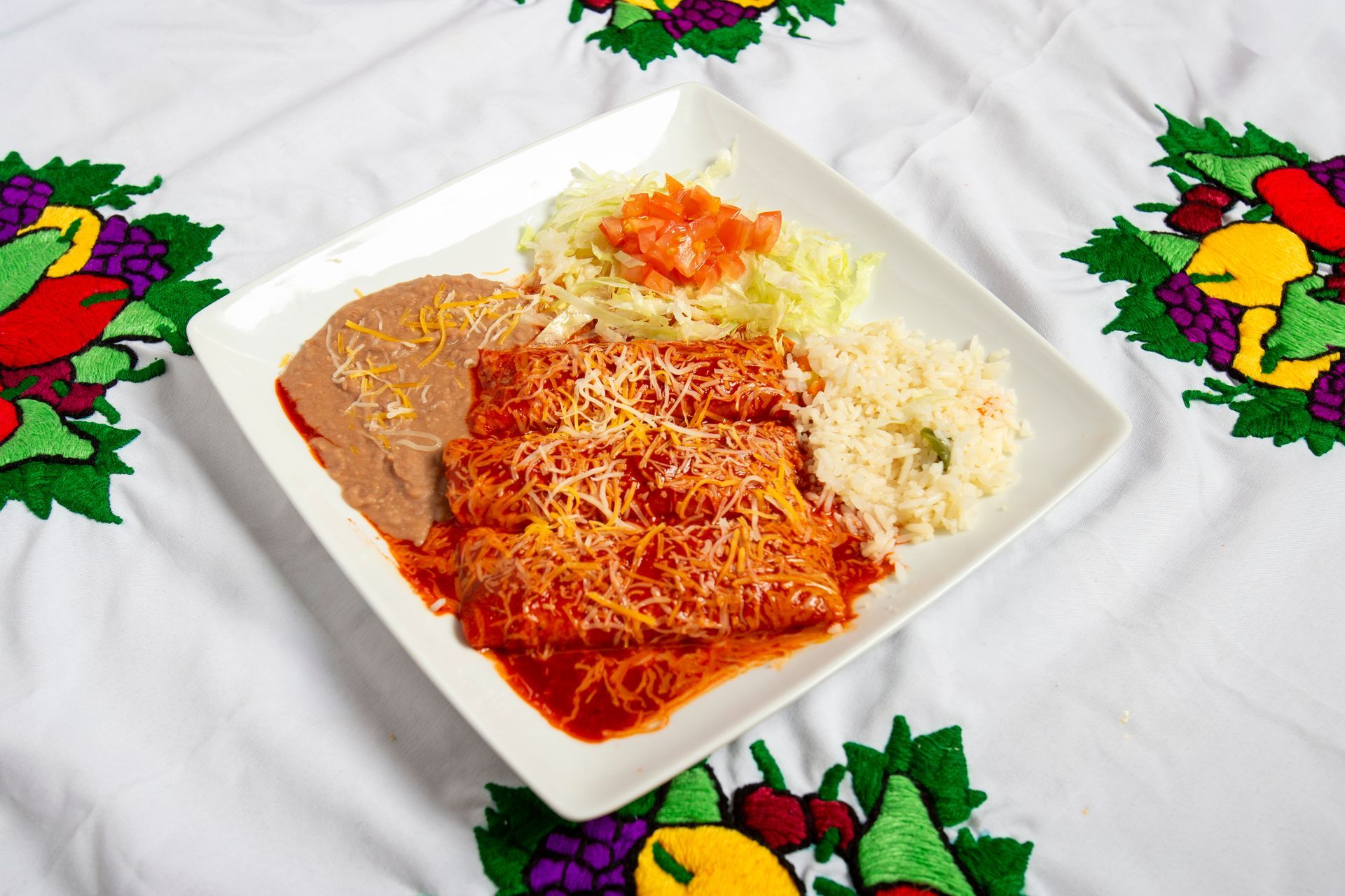 Plate of enchiladas, beans, rice, and cabbage with salsa, on a decorative tablecloth.