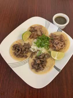 Four tacos on a white plate, topped with meat, cilantro, and onion, served with lime and a small cup of salsa.