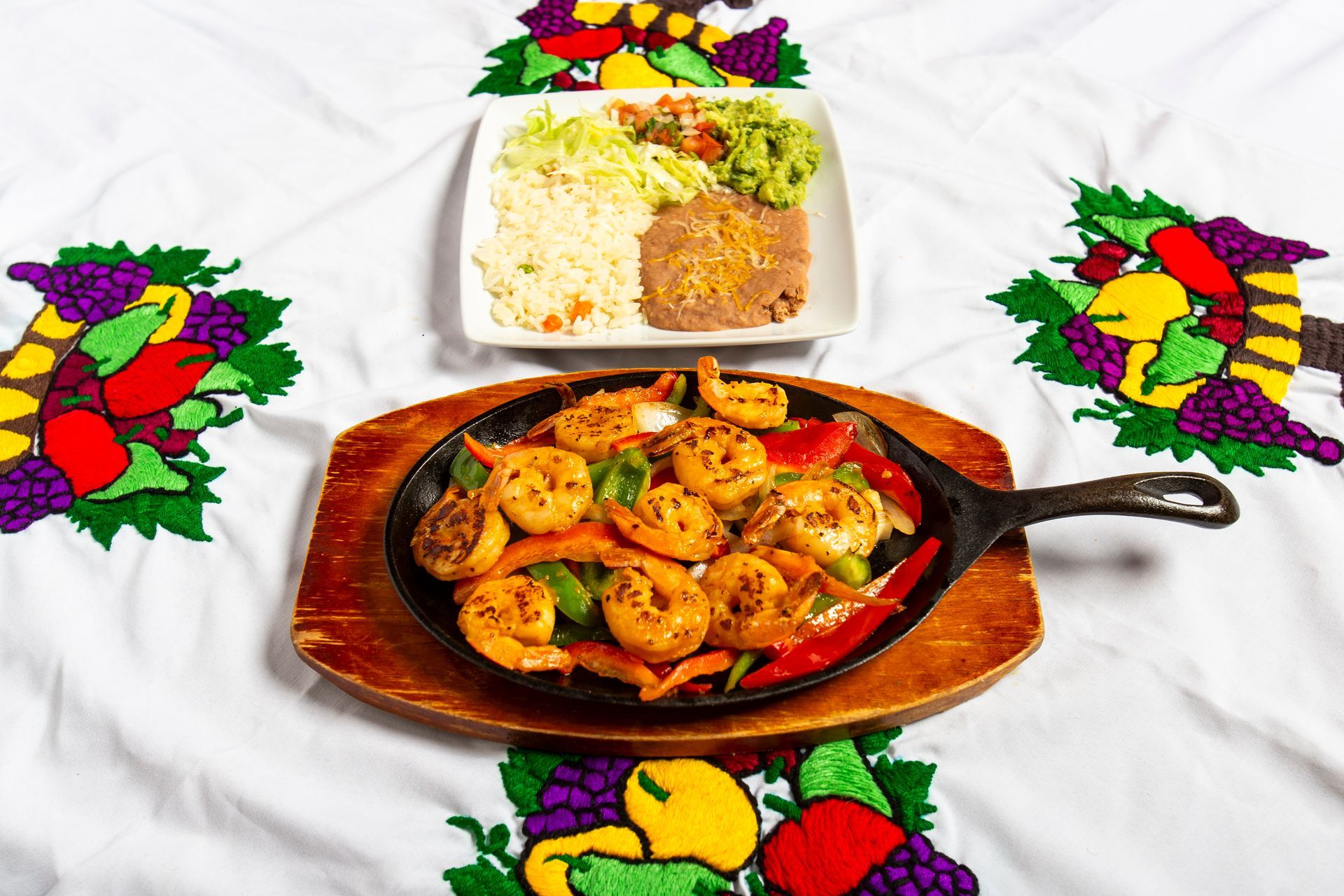 Shrimp fajitas sizzling in a cast iron pan, served with rice, beans, and guacamole on a white cloth with fruit embroidery.