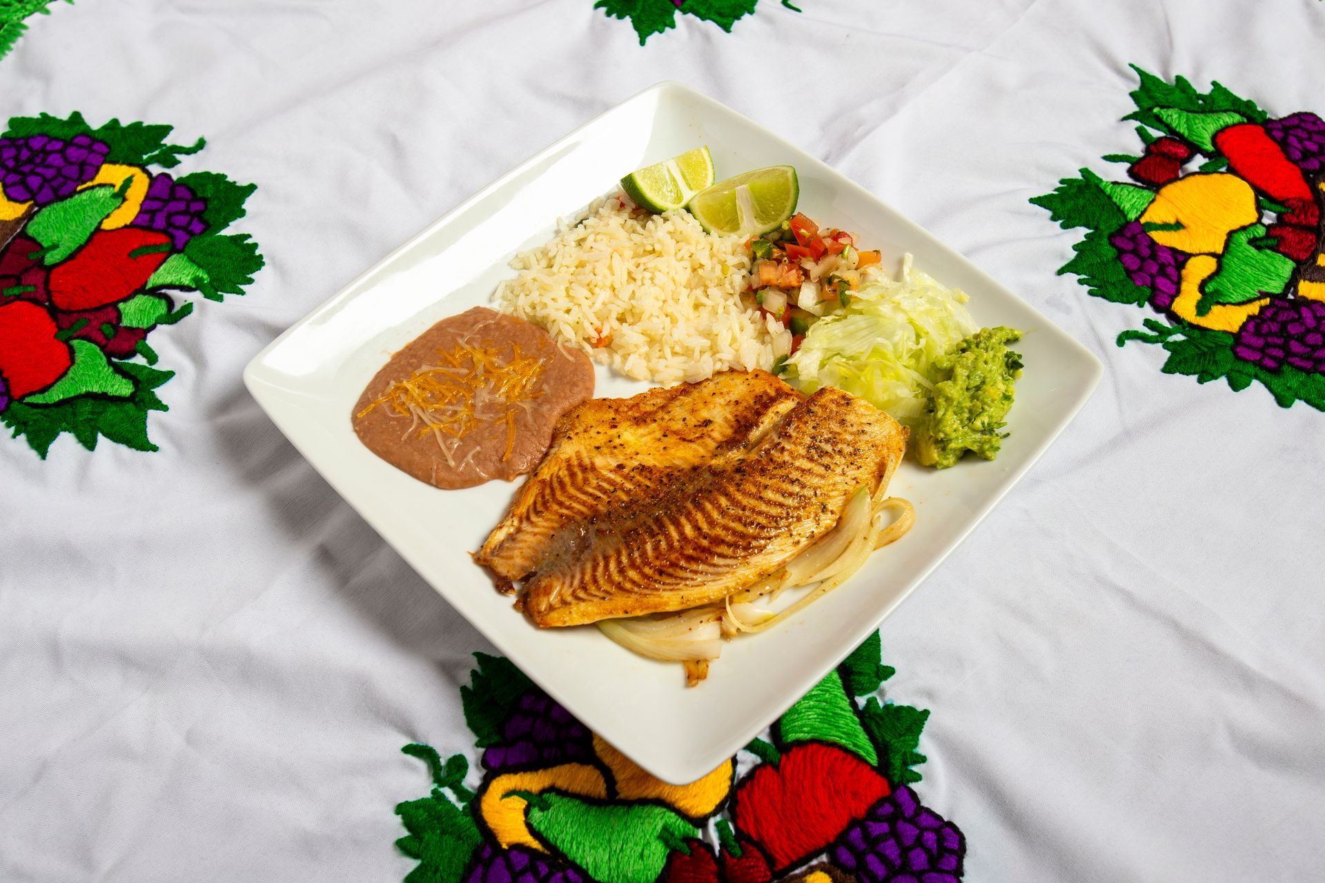Plate of Mexican food: grilled fish, rice, beans, salsa, guacamole, and lime on embroidered tablecloth.