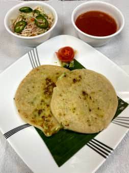 Two pupusas with toppings, a bowl of slaw, and a bowl of red sauce on a white plate.