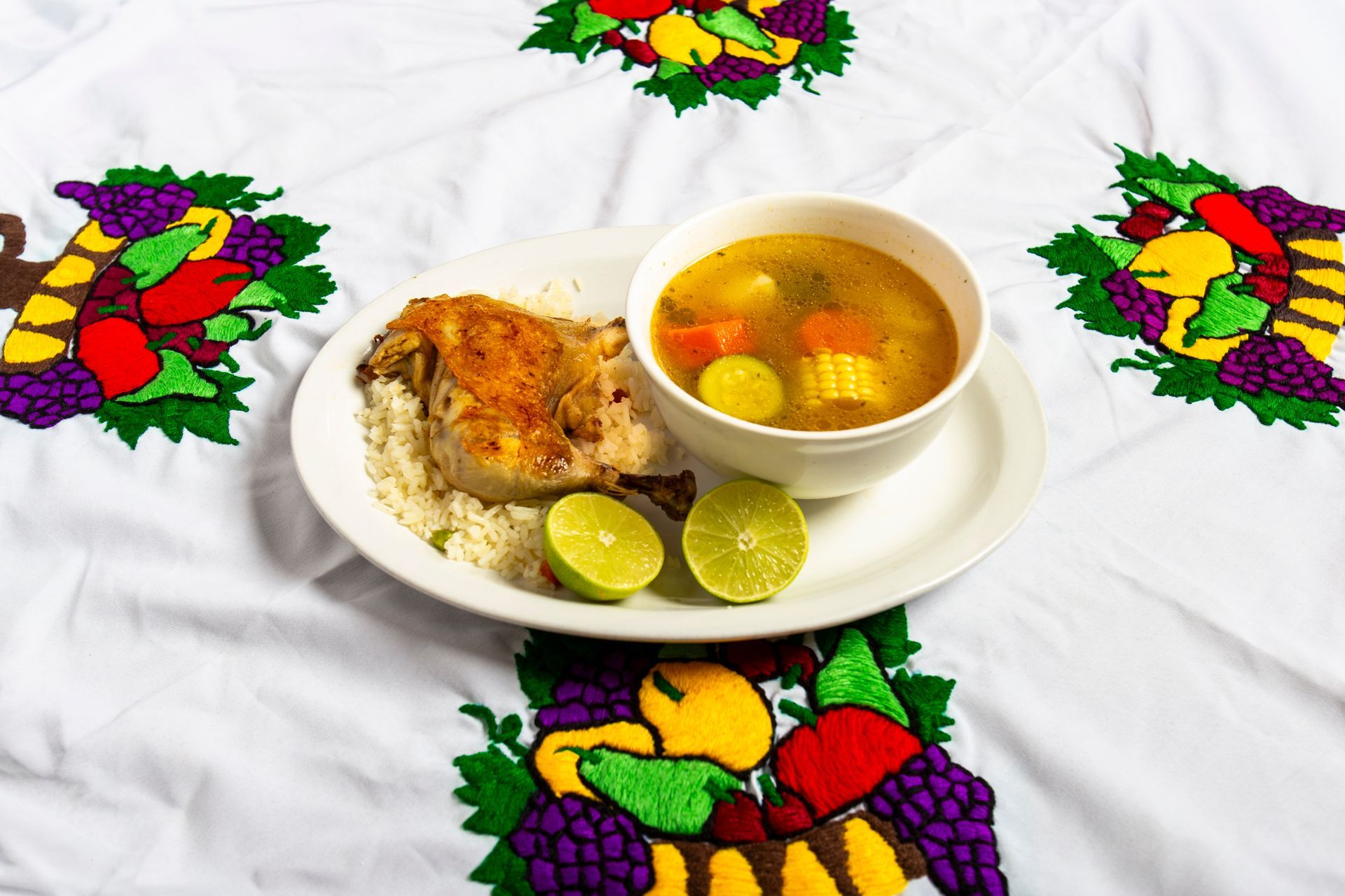 Plate with chicken and rice, soup bowl with vegetables, and lime halves on embroidered tablecloth.