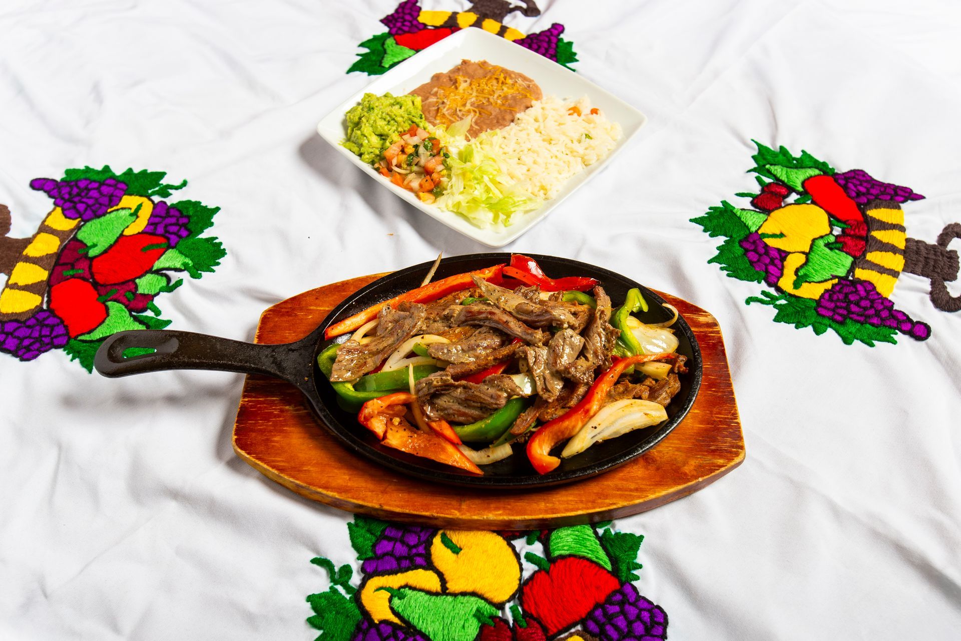 Fajitas in a cast iron pan with a side dish on a table with embroidered decorations.
