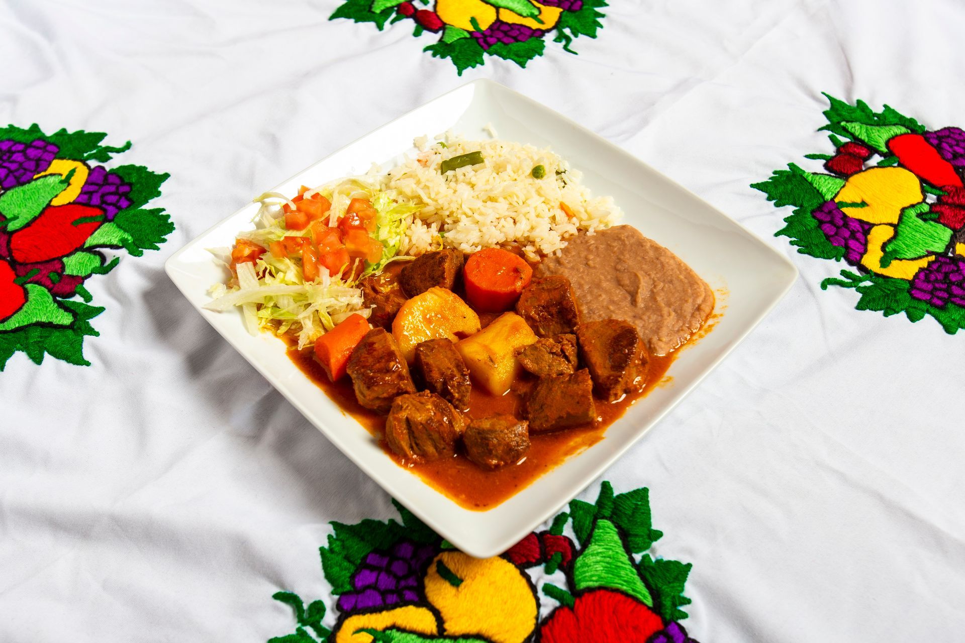 A plate with stewed meat, rice, beans, and slaw on an embroidered tablecloth.