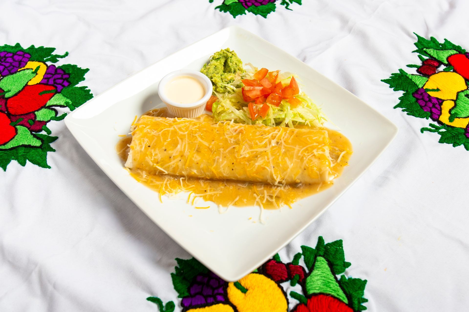 Burrito covered in sauce, on a white plate with guacamole, tomatoes, and sour cream.