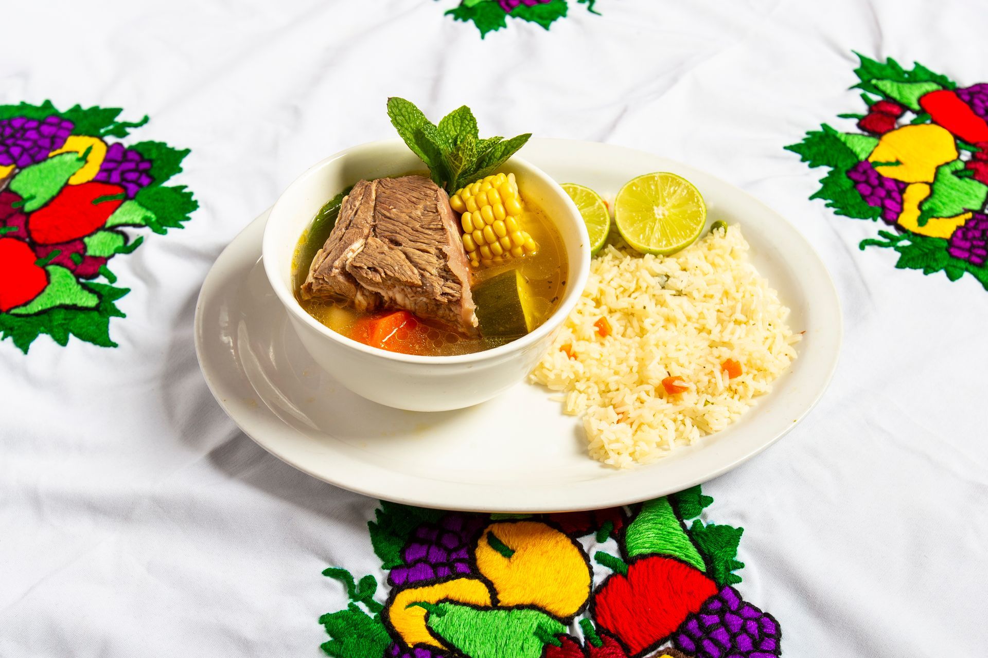 Soup with meat and vegetables, corn, side of rice, lime, and mint garnish on a white plate.