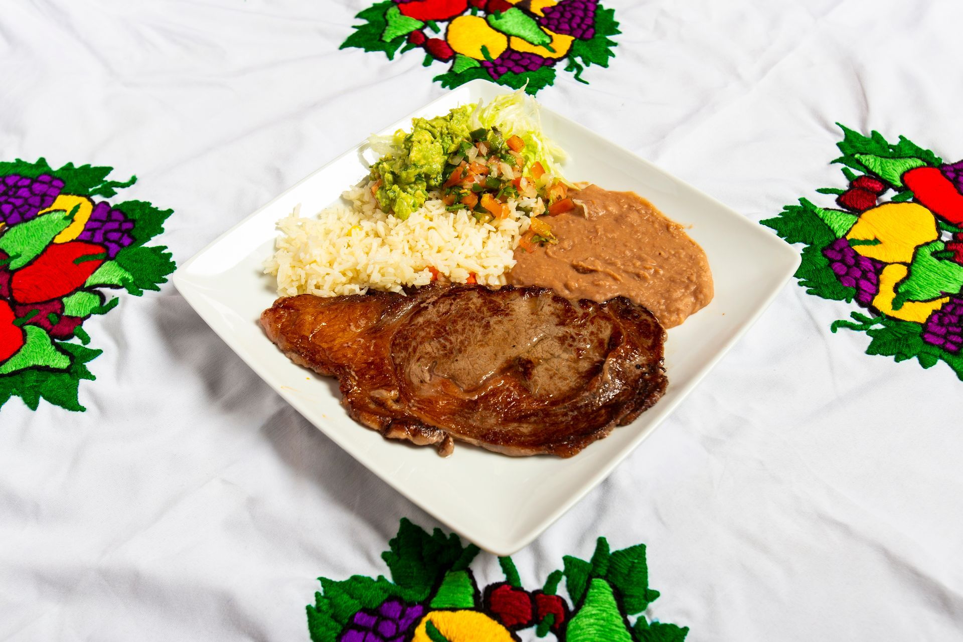 Plate of steak, rice, refried beans, and guacamole on embroidered tablecloth.