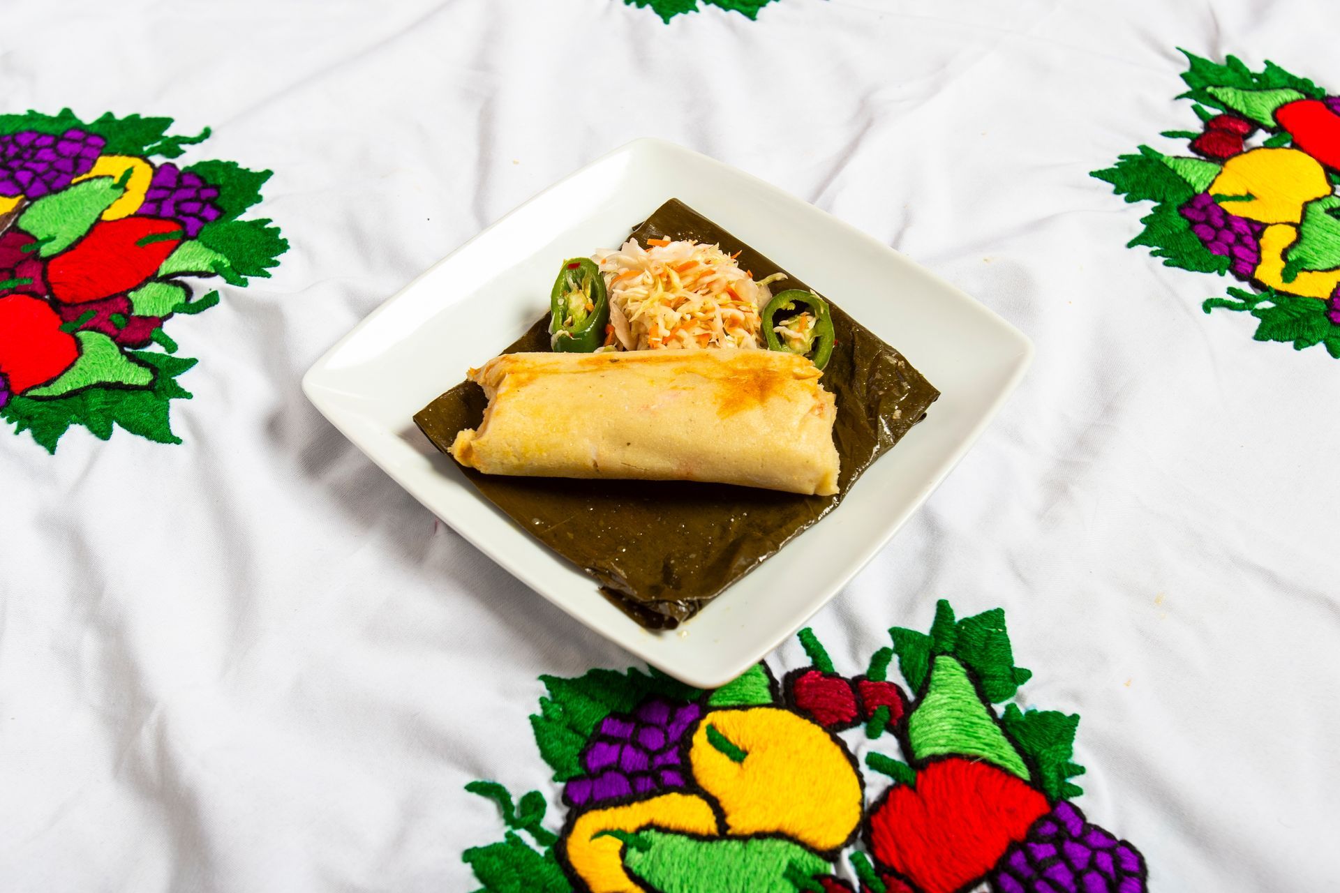 Tamale with rice and jalapeños on a white square plate, set on a floral embroidered tablecloth.