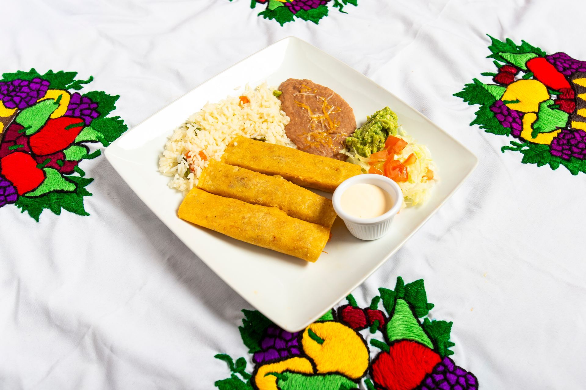 Plate of Mexican food: rice, beans, taquitos, guacamole, and sour cream on a white, embroidered tablecloth.
