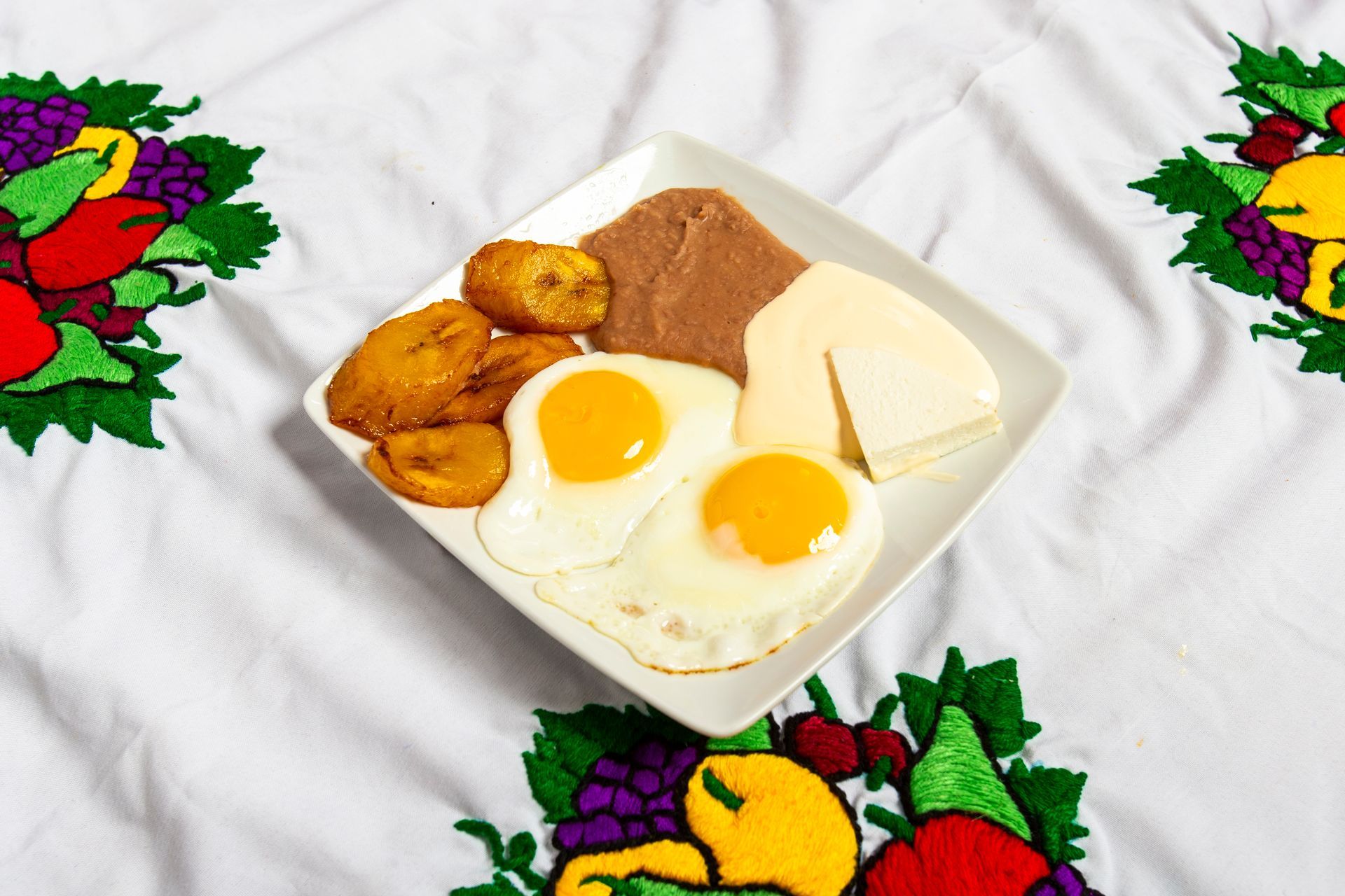 Breakfast plate: fried eggs, plantains, refried beans, cheese, on embroidered tablecloth.