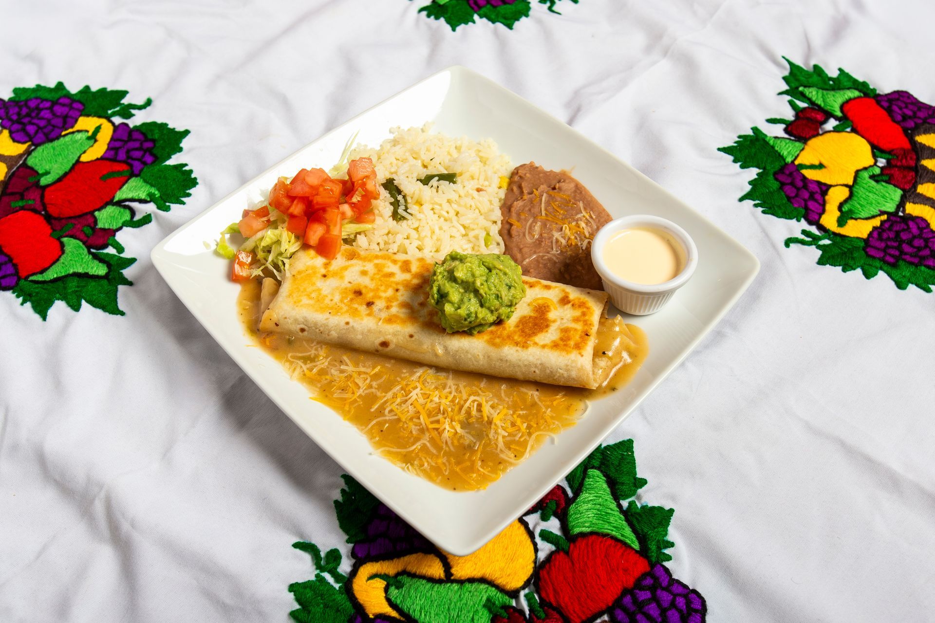 Plate of Mexican food: burrito, rice, beans, salsa, guacamole, and sauce, on a white embroidered tablecloth.