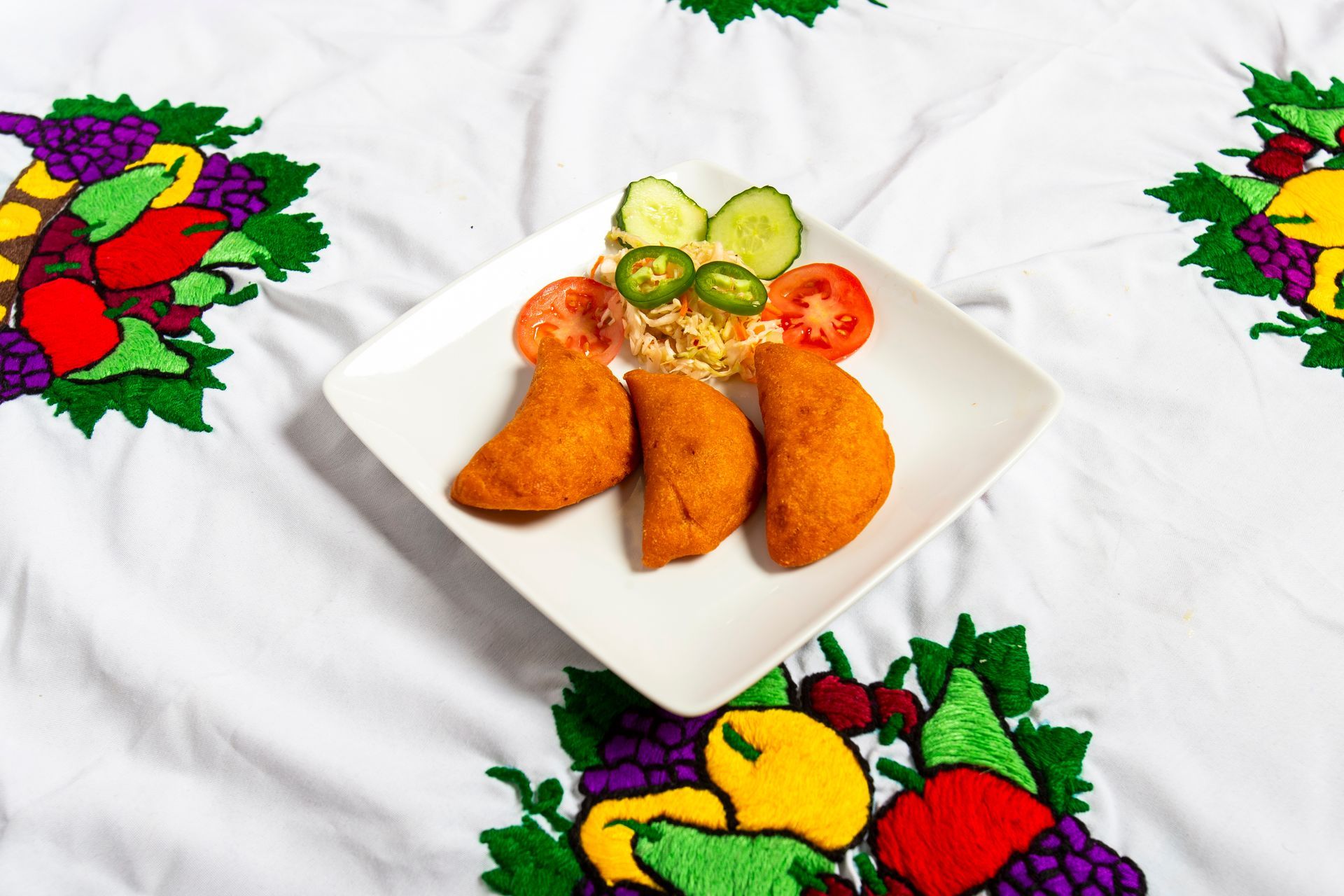 Three fried pastries on a white plate with tomatoes, cucumber, jalapeños, and slaw on a cloth with fruit embroidery.