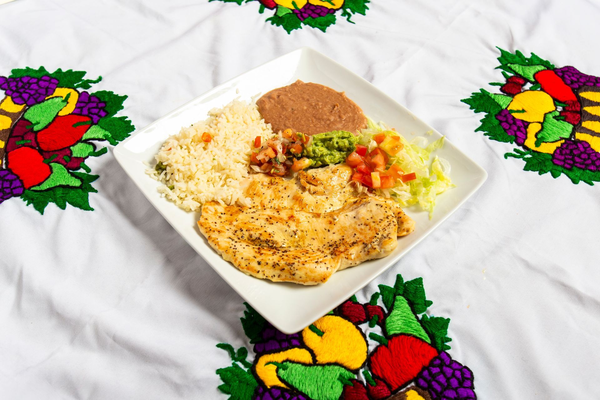 Plate with grilled chicken, rice, beans, guacamole, and salsa on embroidered tablecloth.