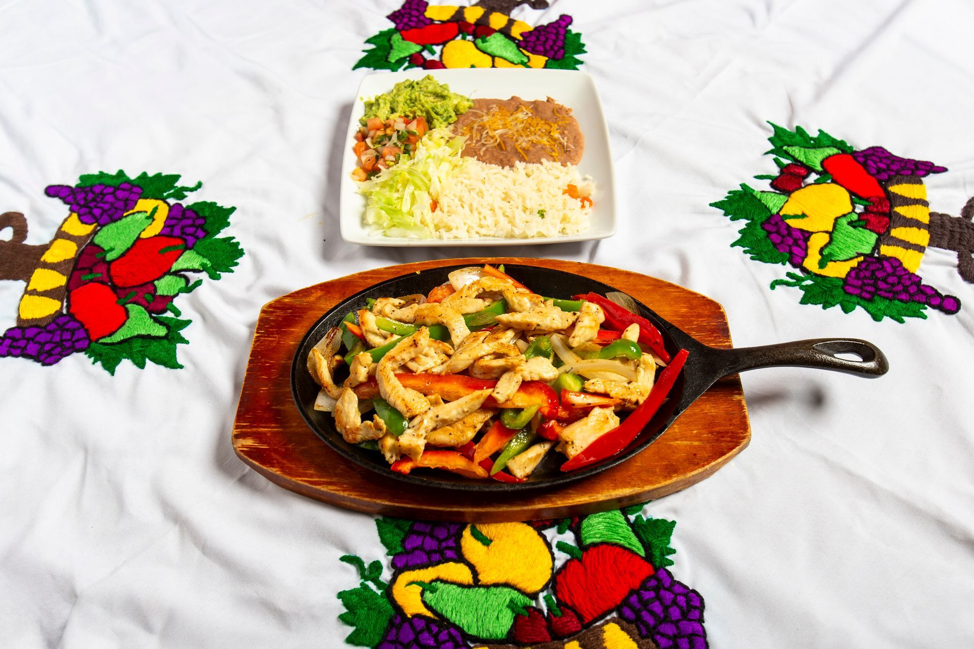 Chicken fajitas in a skillet with side of rice, beans, and guacamole, on a decorative tablecloth.