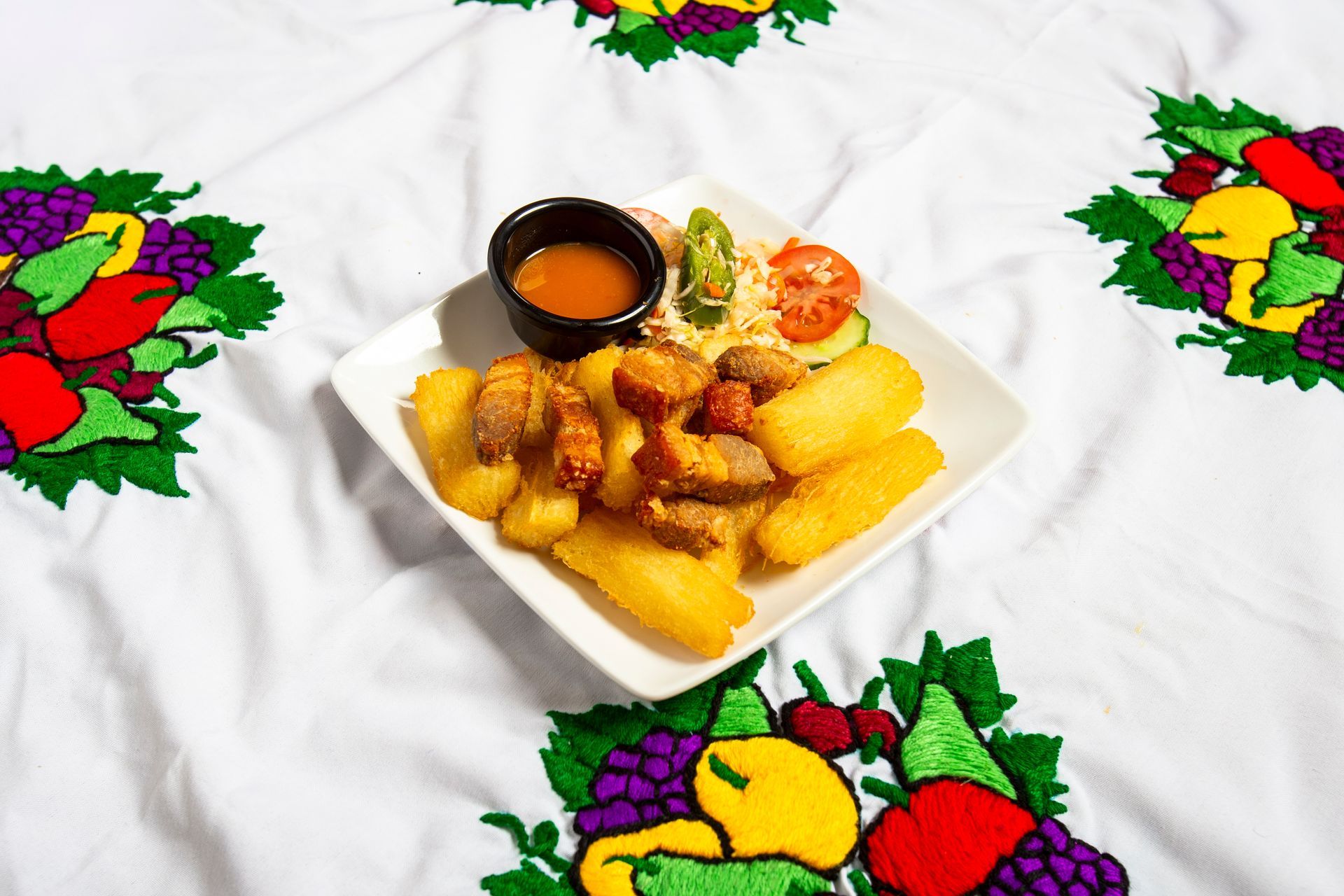 Plate of fried food with a small dish of sauce and side salad on a tablecloth with colorful fruit embroidery.