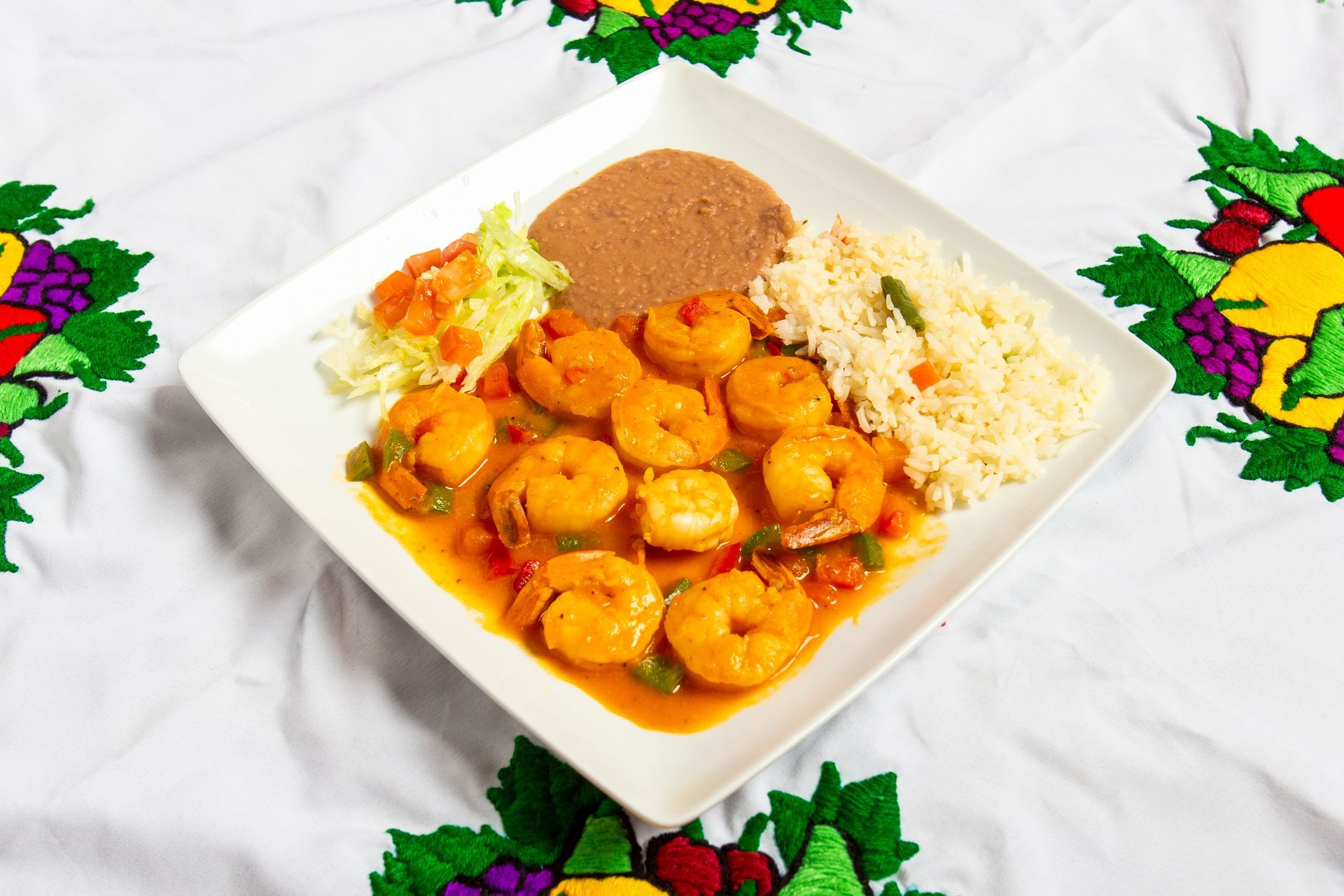 Shrimp dish on white plate with rice, beans, and salad; white tablecloth with colorful embroidery.