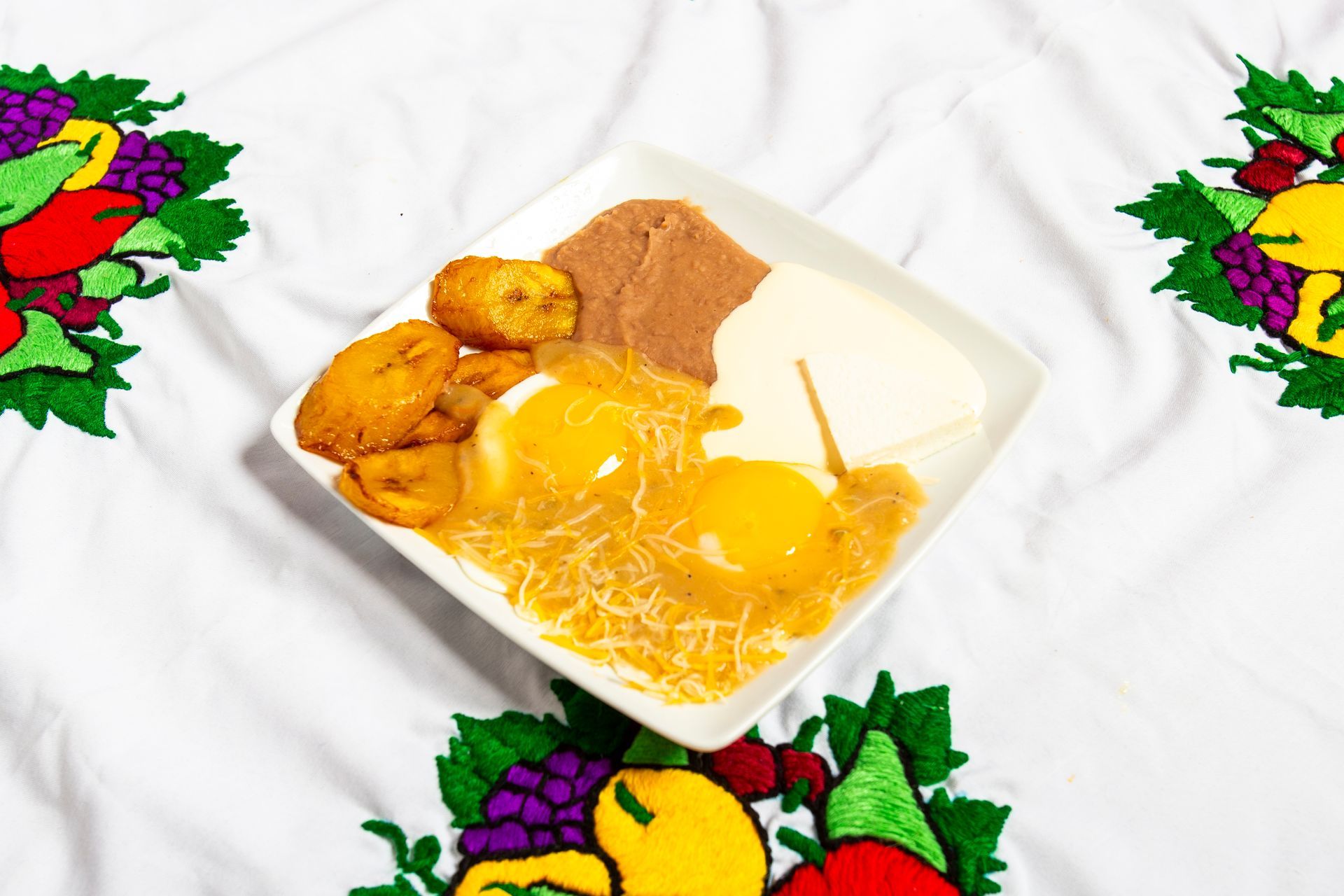Plate of fried eggs, beans, cheese, and plantains on a white embroidered tablecloth.