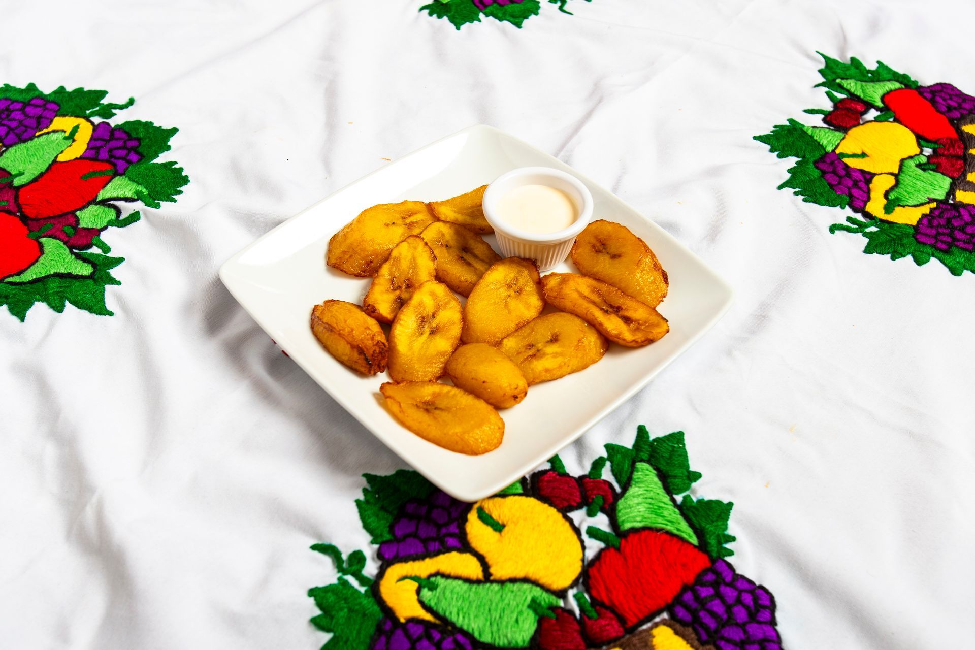 Fried plantains on a white plate with sauce, atop a white tablecloth with embroidered fruit.