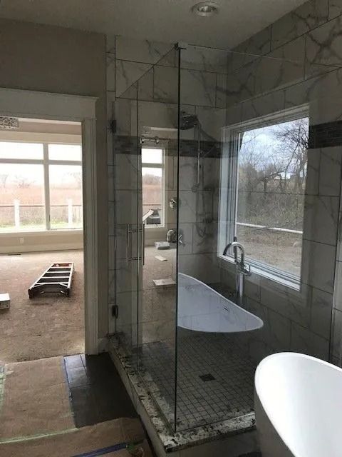 A modern bathroom with a glass shower, a white bathtub, and large windows with an outdoor view.
