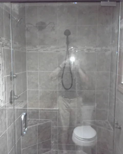 Shower stall with glass door, tiled walls, and a toilet; a person is reflected in the glass.