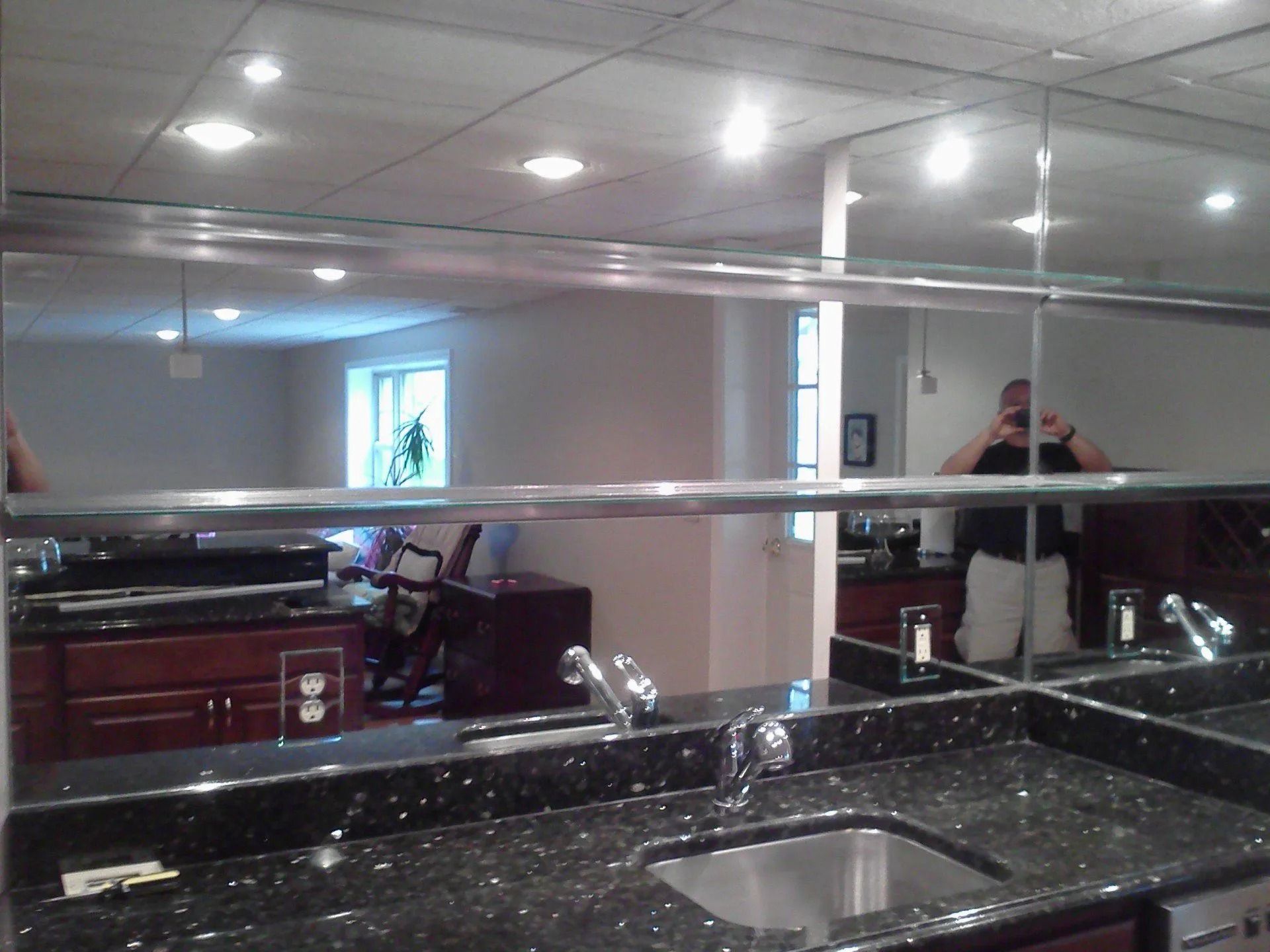 Bar area with a granite countertop, sink, and mirrored backsplash reflecting the room and a person taking a photo.