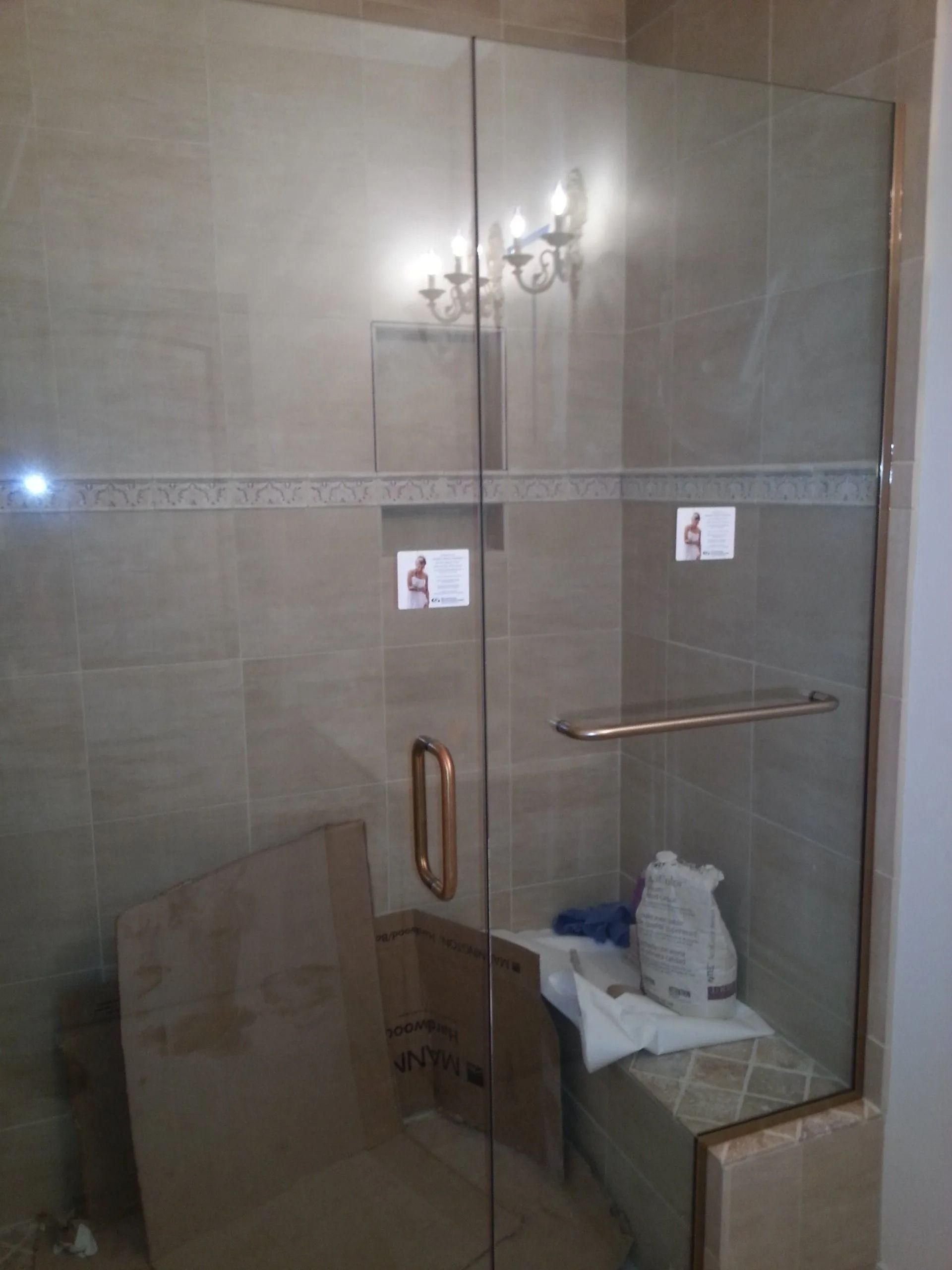 Glass shower enclosure with gold-toned hardware, on tan tiled wall. A bench is inside, with some items.