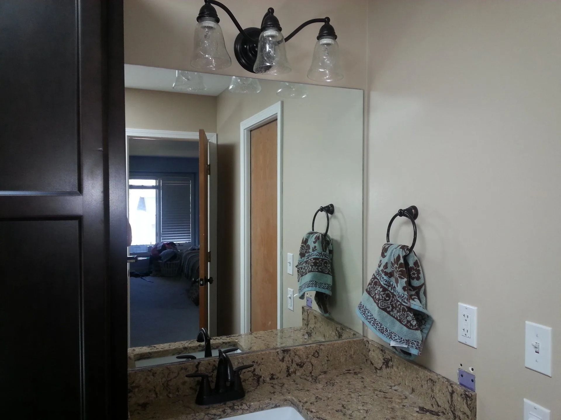 Bathroom with mirror, lights, and granite countertop reflecting a glimpse of the hallway. Two towels hang.