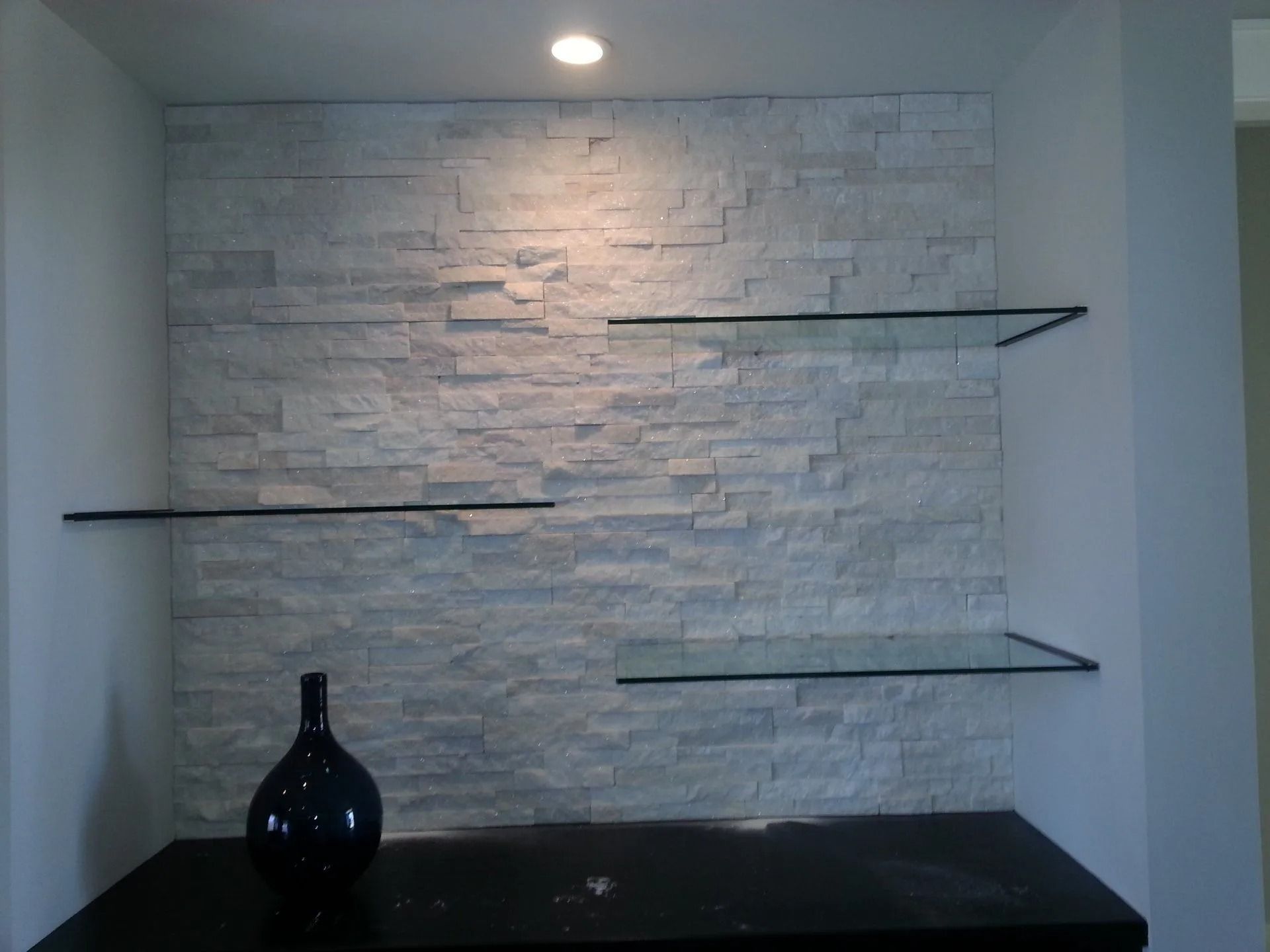 White stone wall with inset glass shelves, lit by a recessed light. A dark blue vase sits on a black surface below.