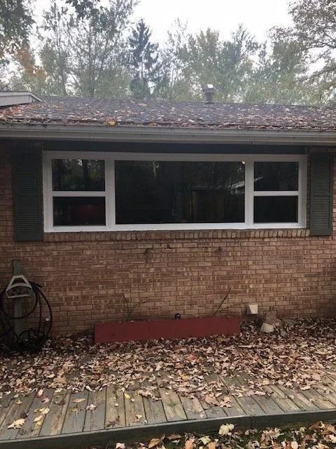 Brick house with large white-framed window. Green shutters flank the window. Fallen leaves on a wooden deck.