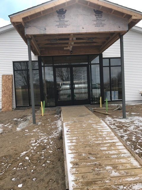 Entrance of a building with a wooden ramp. Glass doors and windows under a covered overhang. Construction site with some snow.