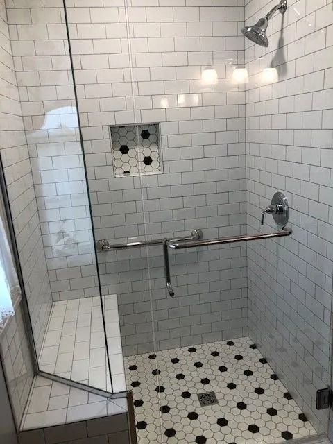 Shower with white subway tile, a glass door, built-in seat, and grab bar, featuring a black and white hex tile floor.