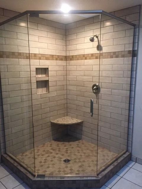 Corner shower stall with glass doors, tiled walls, built-in seat and shelf. Beige and brown tones.