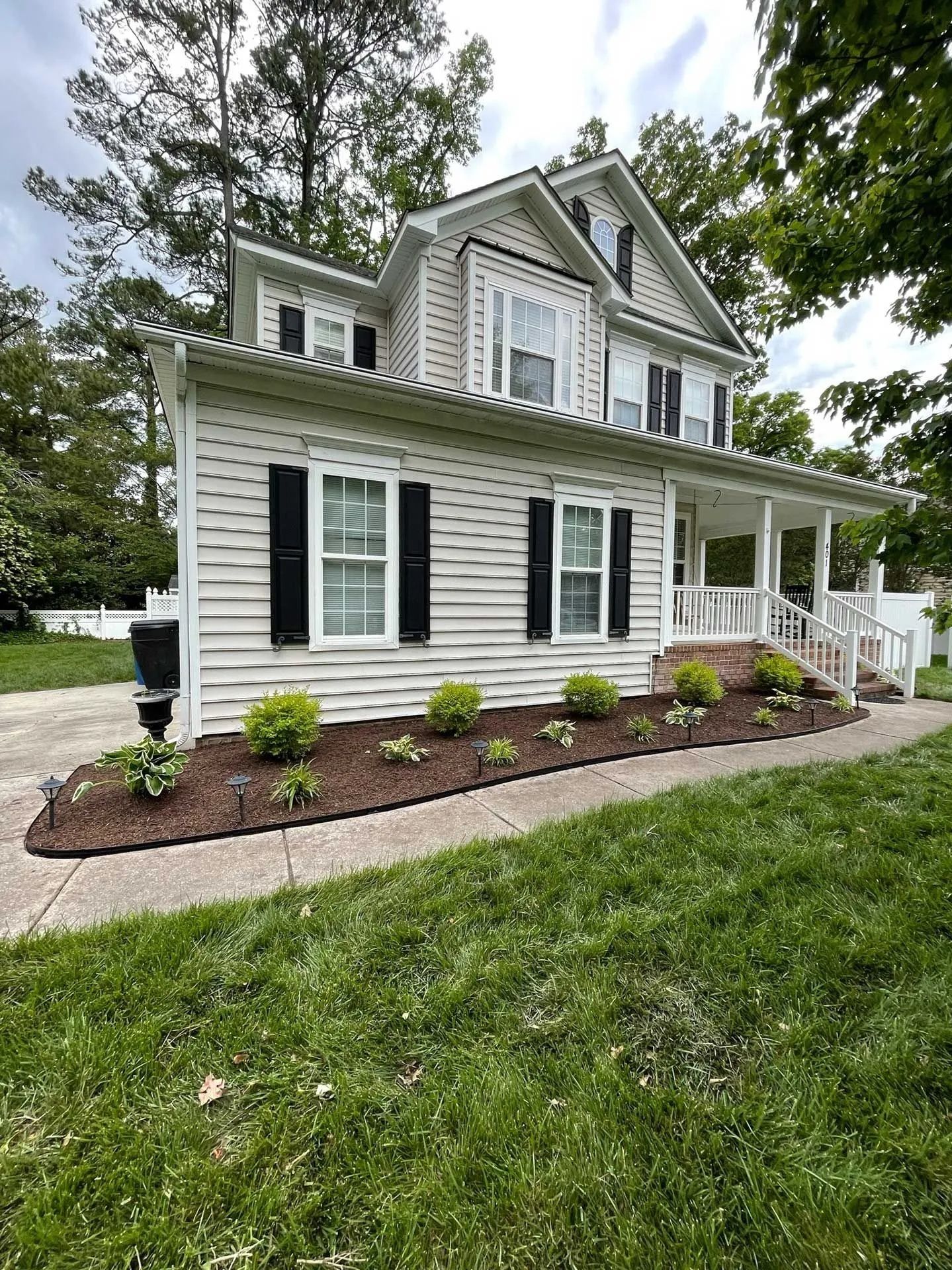 White two-story house with black shutters, front porch, and landscaped yard surrounded by trees