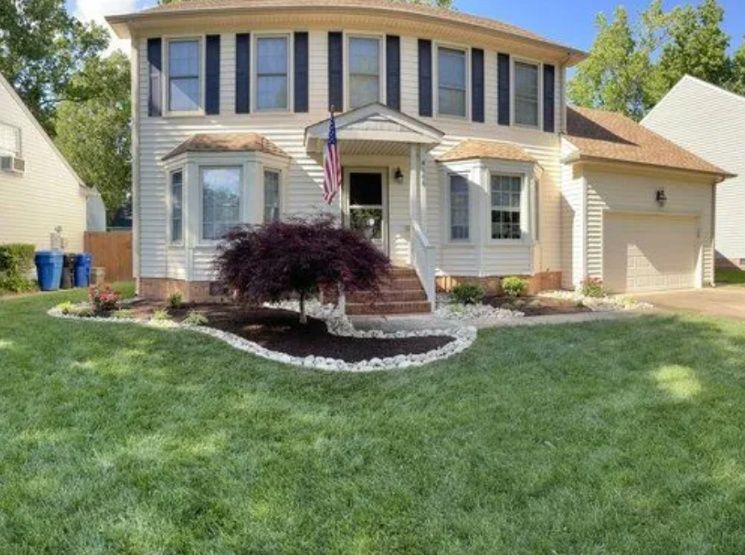 Two-story beige house with a front lawn, porch, and a small tree in the yard