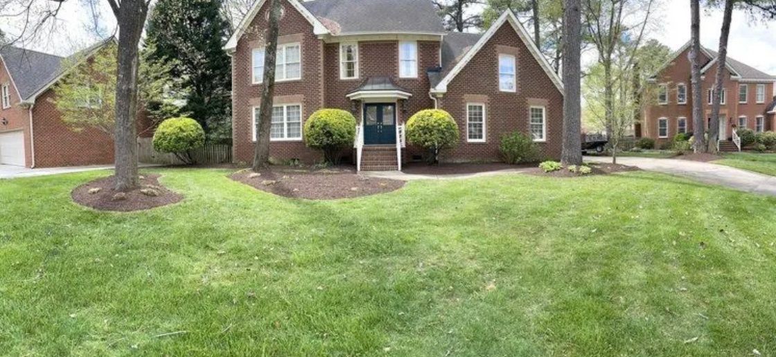 Brick suburban house with manicured lawn, shrubs, and a driveway under overcast skies