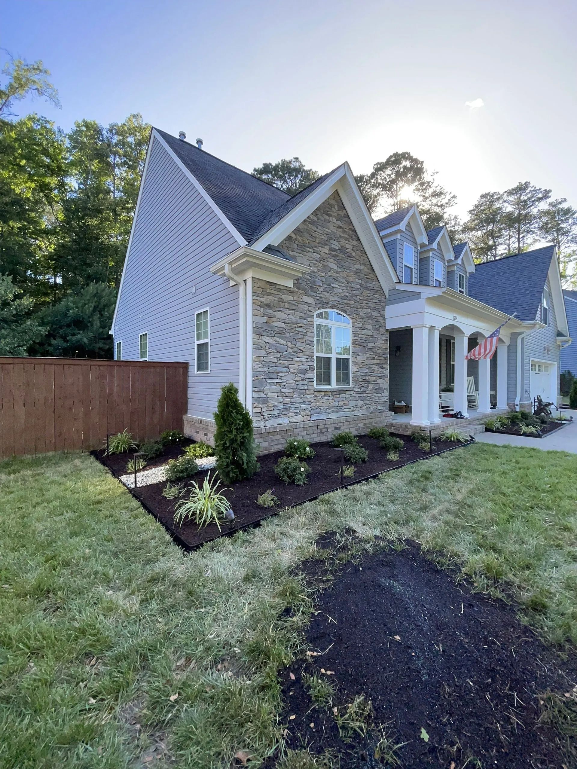 Stone house with steep roof, white trim, and flower beds beside a lawn at sunset