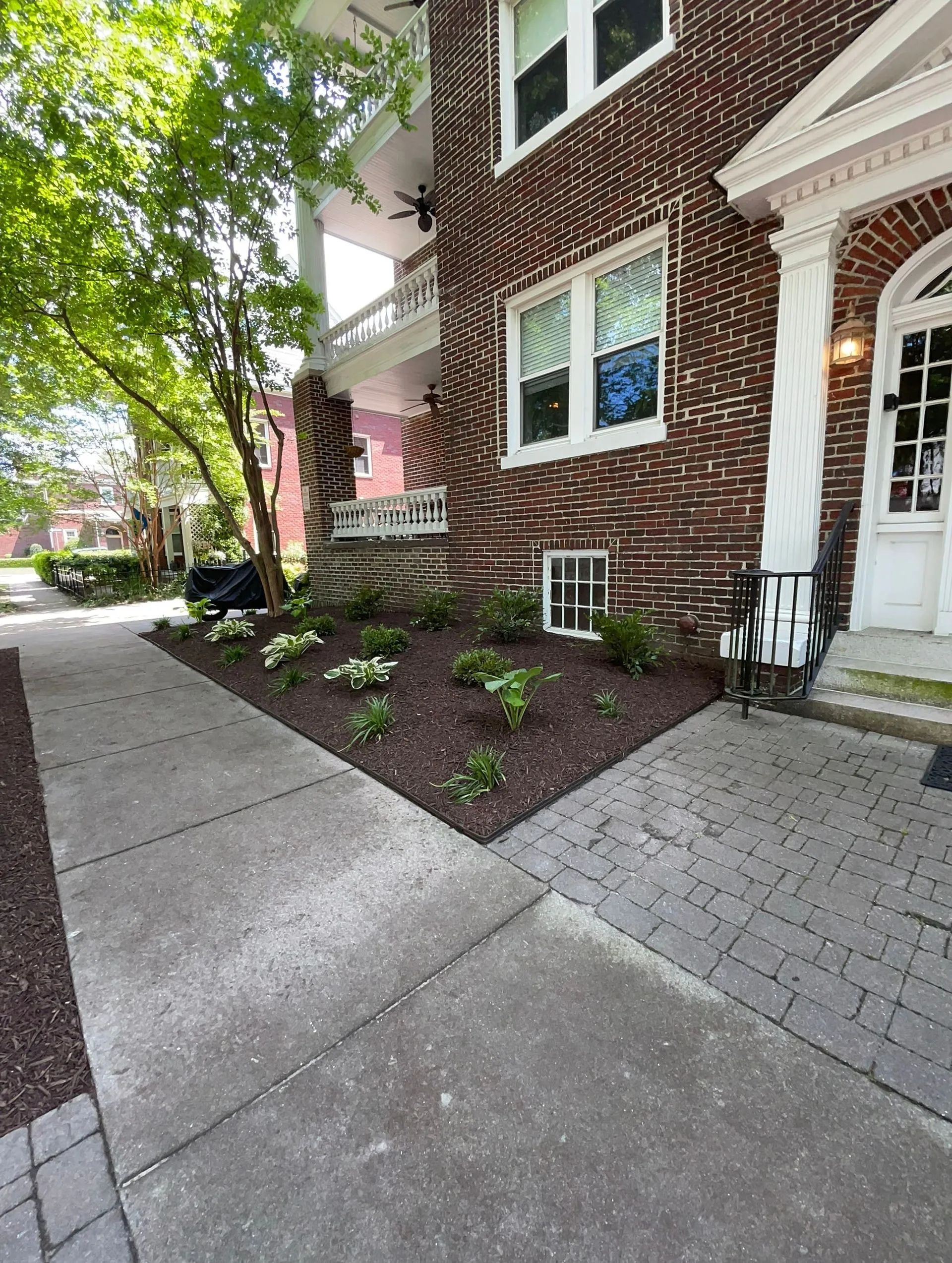 Brick apartment entrance with white door, sidewalk, landscaping, and trees along the walkway