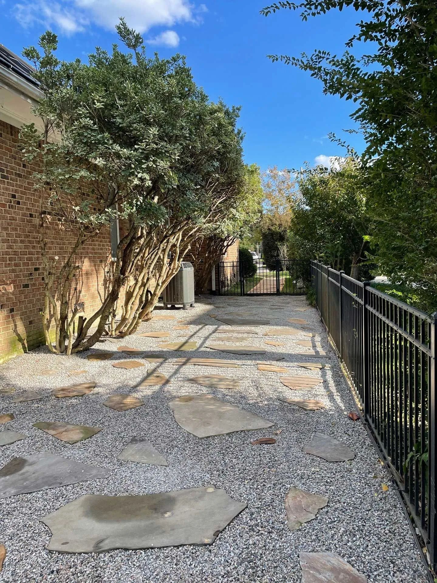 Stone walkway beside brick wall and black fence, lined with trees under a bright blue sky.