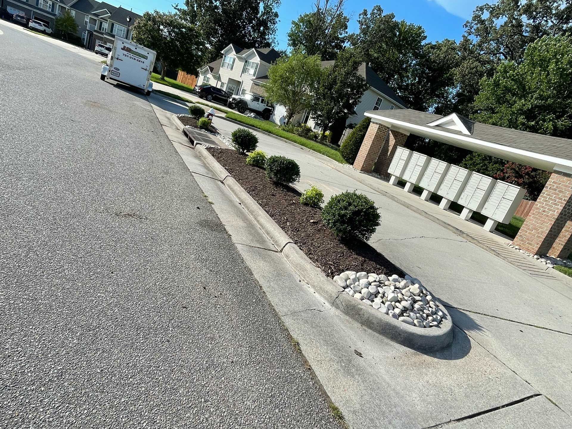 Residential street with landscaped curb, mailbox shelter, and gravel road on a sunny day