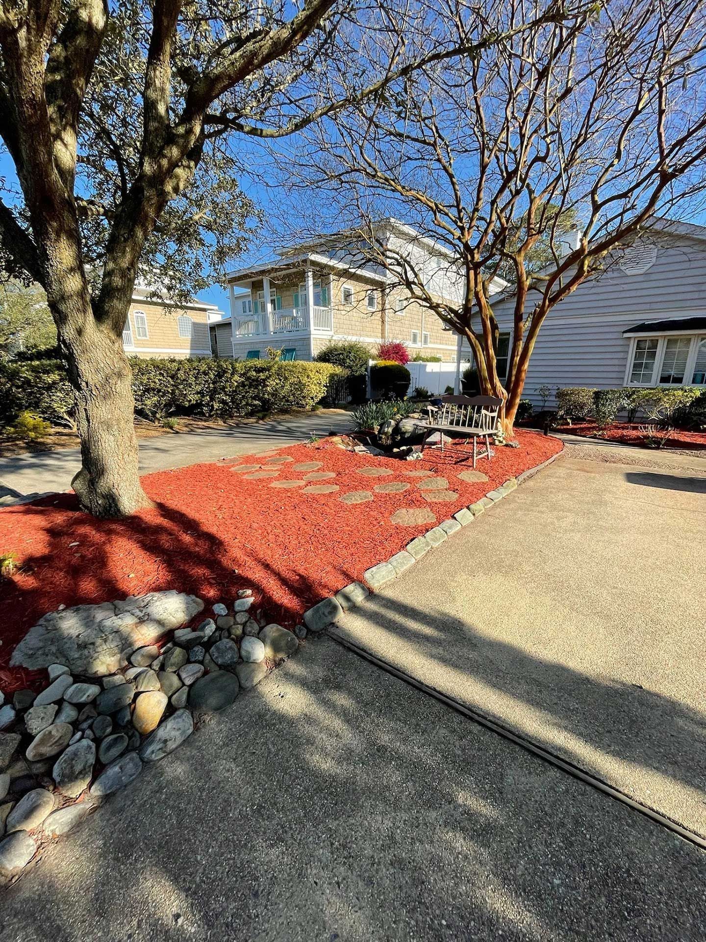 Front yard with red mulch, trees, and a curved concrete driveway beside a house under a clear blue sky