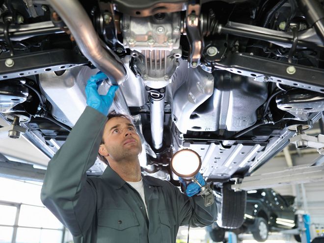 Mechanic examines car undercarriage with a flashlight in a garage.