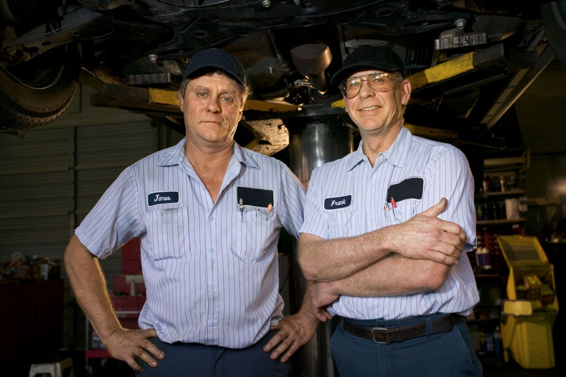 Two mechanics in a garage, one wearing a hat. Both are smiling and looking at the camera.