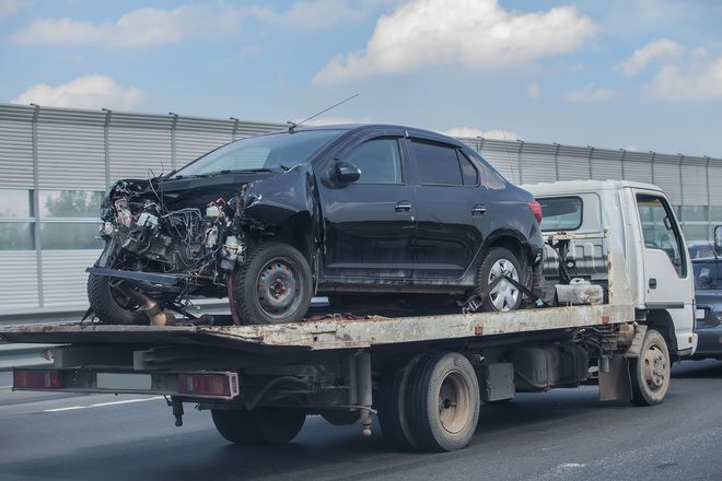 Damaged black car on a flatbed tow truck on a highway.