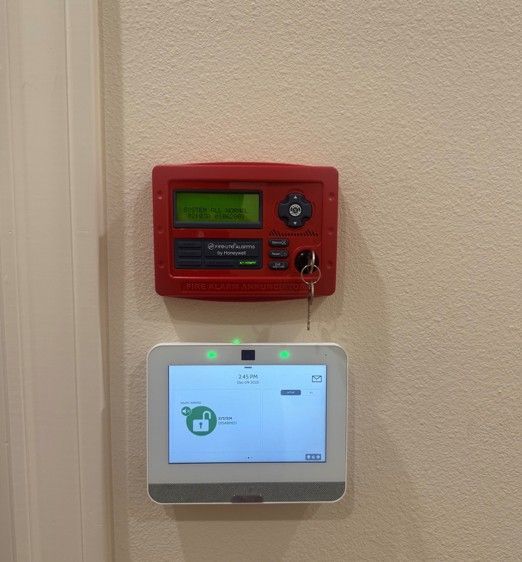 Red wall-mounted alarm panel above a white touchscreen control panel on a beige wall