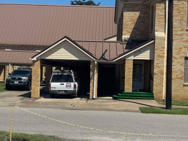 A brick apartment building with covered parking, a white SUV, and a black car in front.