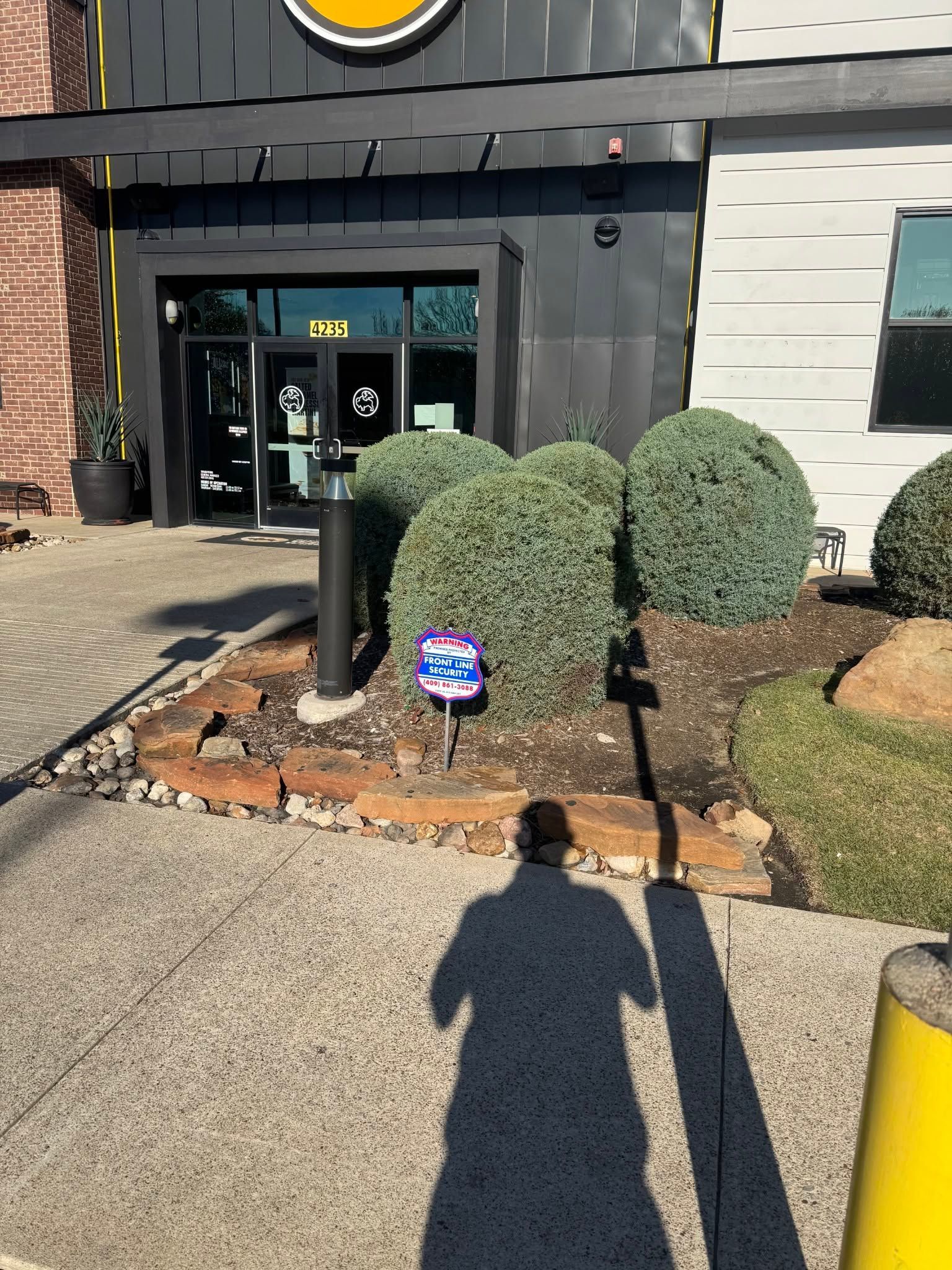 Store entrance with two trimmed shrubs, black bollards, and a person’s shadow on a sunny sidewalk.