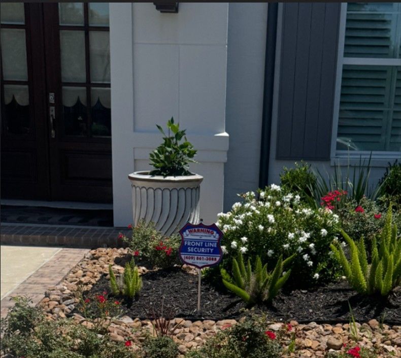 Front garden bed with white flowers, green plants, a potted shrub, and a security sign by a house entrance