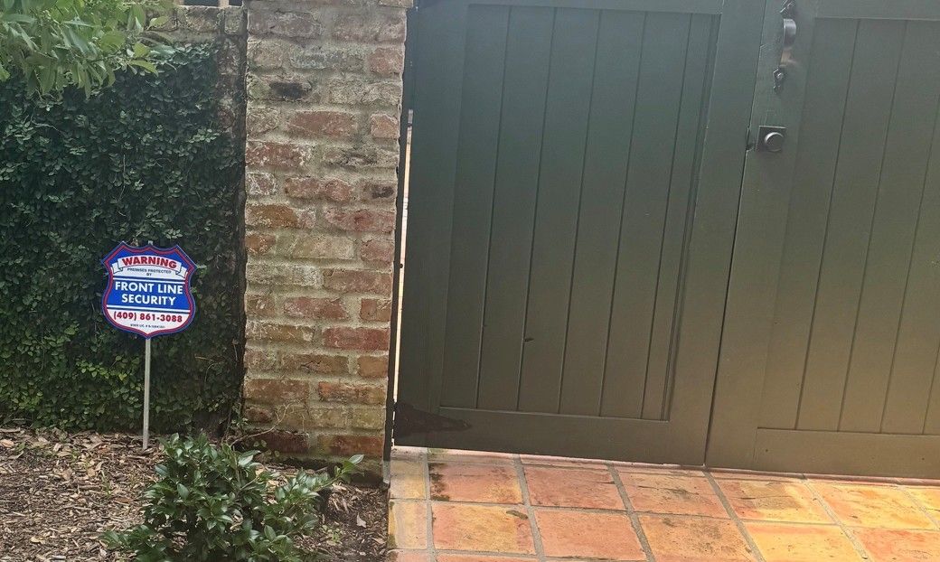 Brick-and-black gate beside a hedge, with a small security sign on a post and tiled walkway in front.